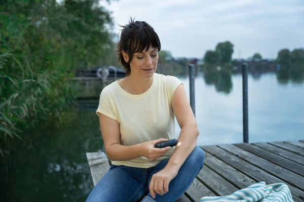 A woman sits on a jetty by the water at dusk and uses the Beurer BR 90.