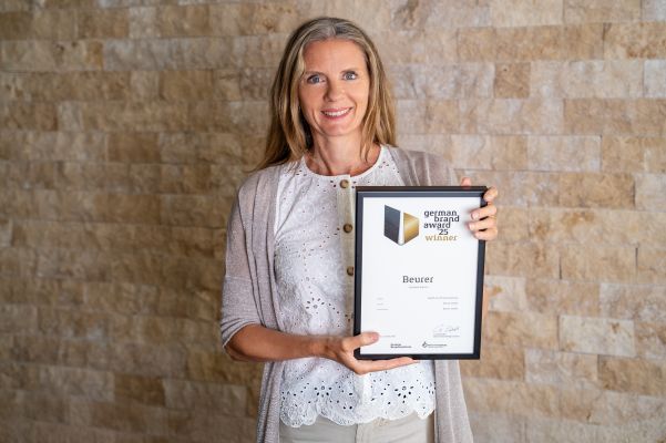 Beurer Director Marketing Kerstin Glanzer stands in front of a stone wall and holds the German Brand Award certificate in her hand.