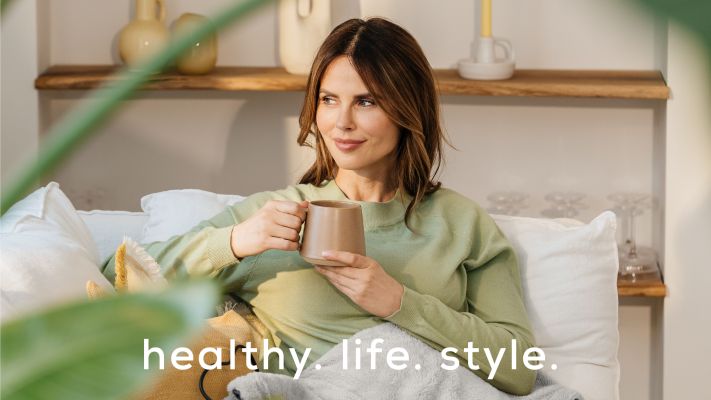 A dark-haired woman sits on a sofa with an electric blanket. Accompanied by the Beurer brand claim healthy. life. style.