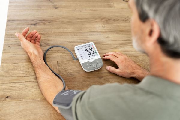 The picture shows an older man sitting at a table and taking a measurement on his upper arm with the Beurer BM 53 blood pressure monitor. 