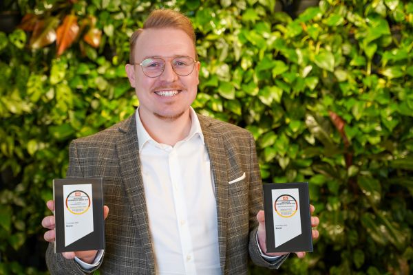 Robin Landgraf, Team Leader Product Management Medical at Beurer, stands in front of a green wall and presents the German Health Award 2025.
