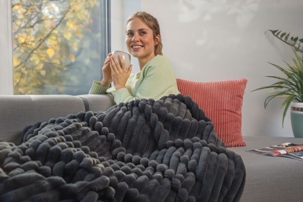 A woman sits by the window with a cup, wrapped in the HD 82 Stockholm thermal blanket.