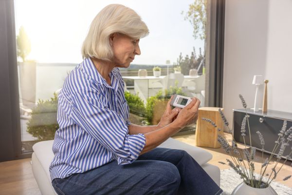 An elderly woman is sitting by the window and using the Beurer ME 95 with her hands.