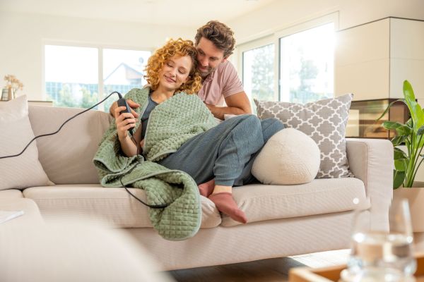 Eine Frau und ein Mann liegen mit der neuen Wärmedecke von Beurer auf dem Sofa