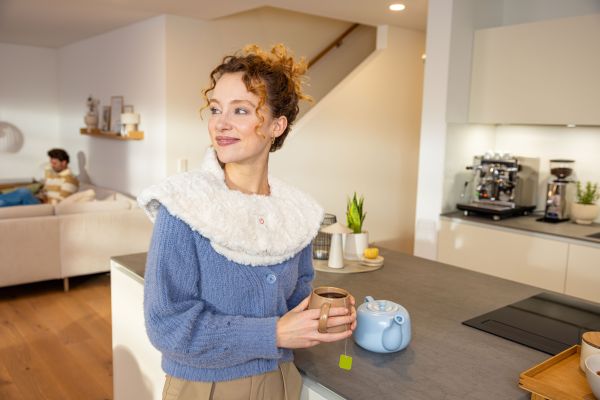 Eine Frau mit Tasse und Nackenheizkissen steht am Tisch und genießt die wohlige Wärme