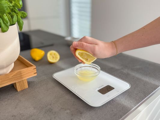 Eine weiße Küchenwaage steht auf einer grauen Aerbeitsfläche und wiegt den Saft einer Zitrone.