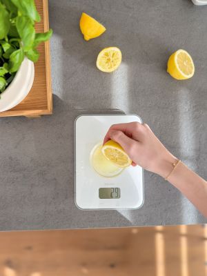 Eine weiße Küchenwaage steht auf einer grauen Aerbeitsfläche und wiegt den Saft einer Zitrone.