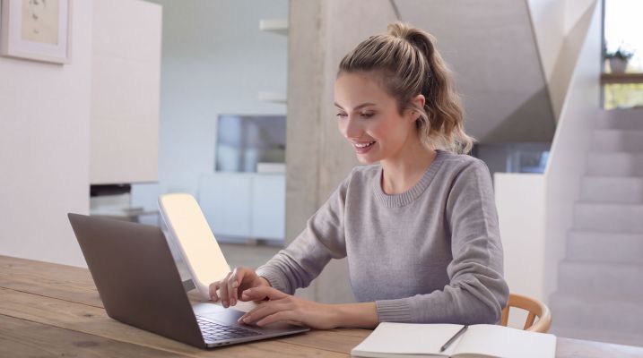 Eine Frau arbeitet am Schriebtisch am Laptip. Im Hintergrund ist die Beurer Tageslichtlampe TL 35 zu sehen- 