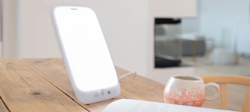 La lampe de luminothérapie Beurer TL 35 est posée dans un salon sur une table en bois à côté d'une tasse et d'un livre