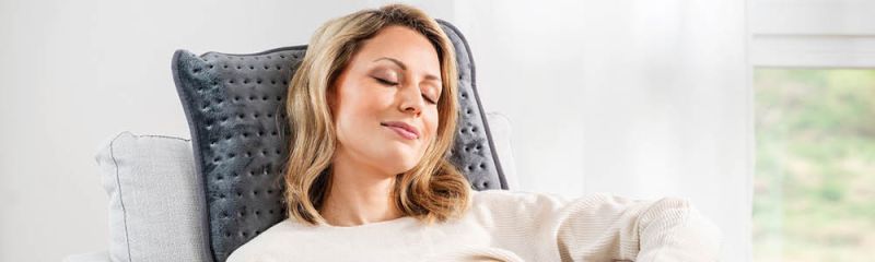 Femme assise dans le fauteuil, appuyée contre le coussin chauffant HK 48 cosy, profite les yeux fermés d'une pause tranquille