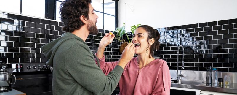 Un couple se tient dans la cuisine, se tendant mutuellement de petites collations et riant
