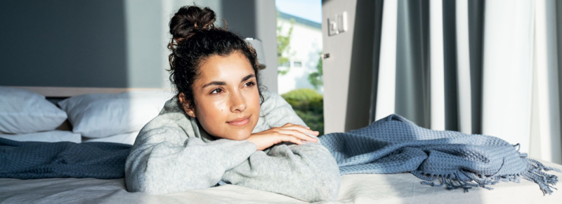 Jeune femme allongée pensivement sur le lit et regardant par la fenêtre, la lumière du jour entre dans la chambre.