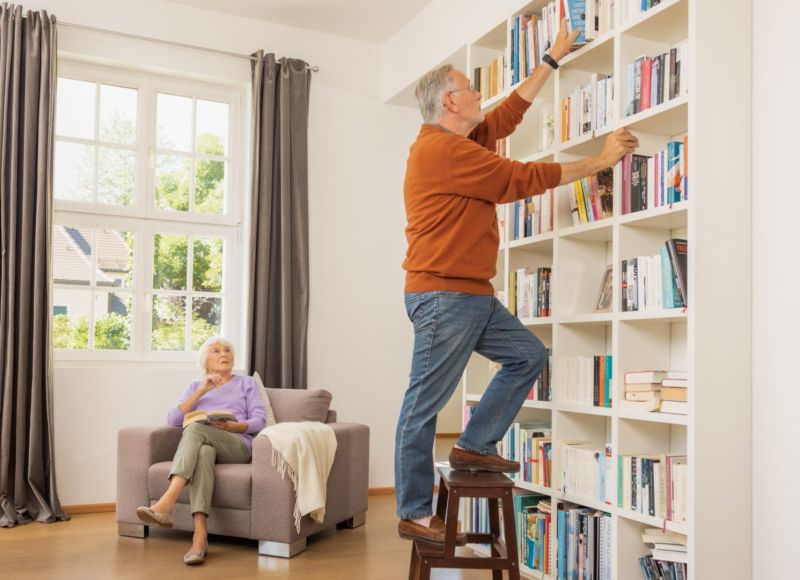 Anziano con braccialetto di emergenza Gardia in piedi su una scaletta di casa e prende un libro, donna seduta su una poltrona