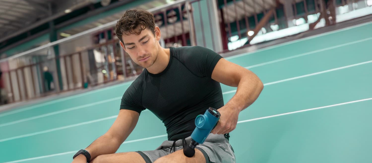 Mann nutzt die Massage Gun AMG 98 BOOM am Oberschenkel auf einer Treppe in einer Turnhalle