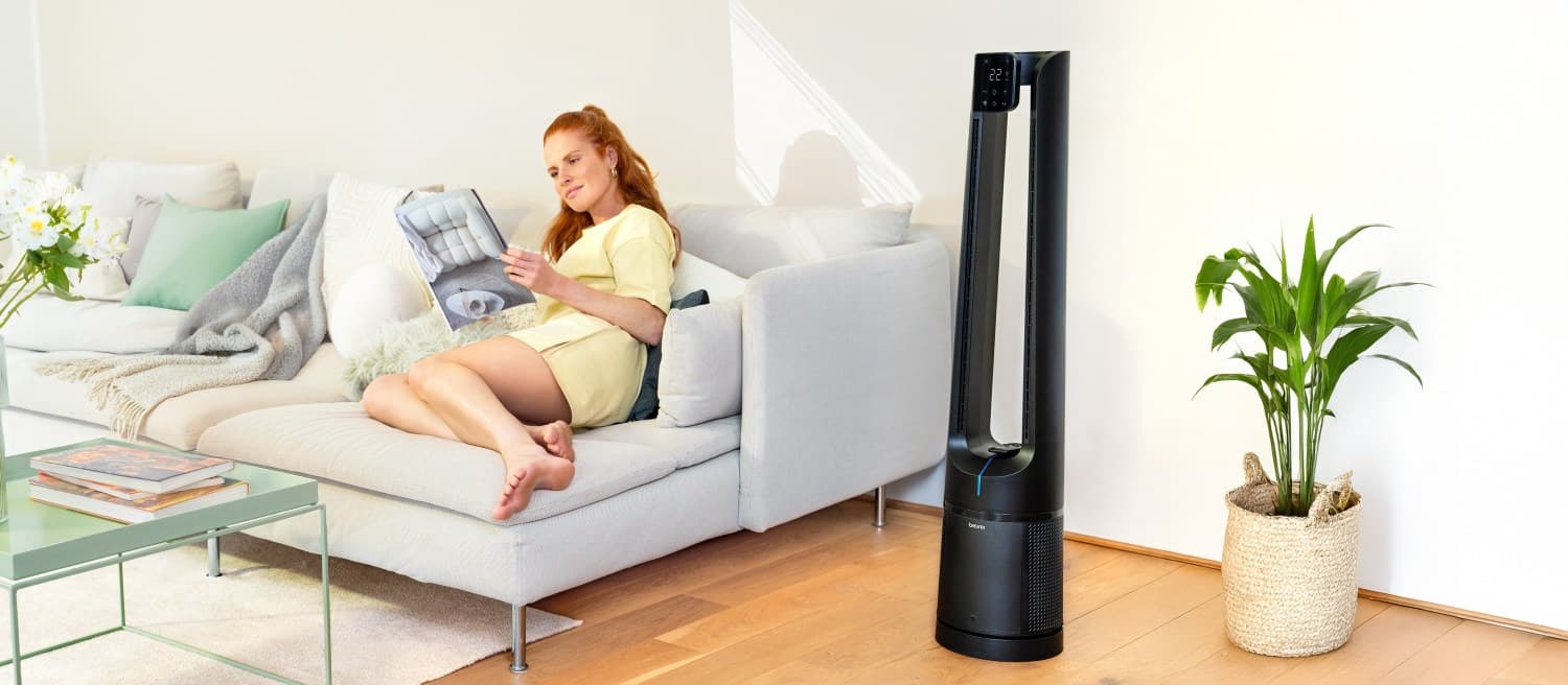 Frau liegt auf einem grauen Sofa und blättert in einer Zeitschrift. Daneben steht der Beurer 2-in-1 Luftreiniger und Ventilator LV 500