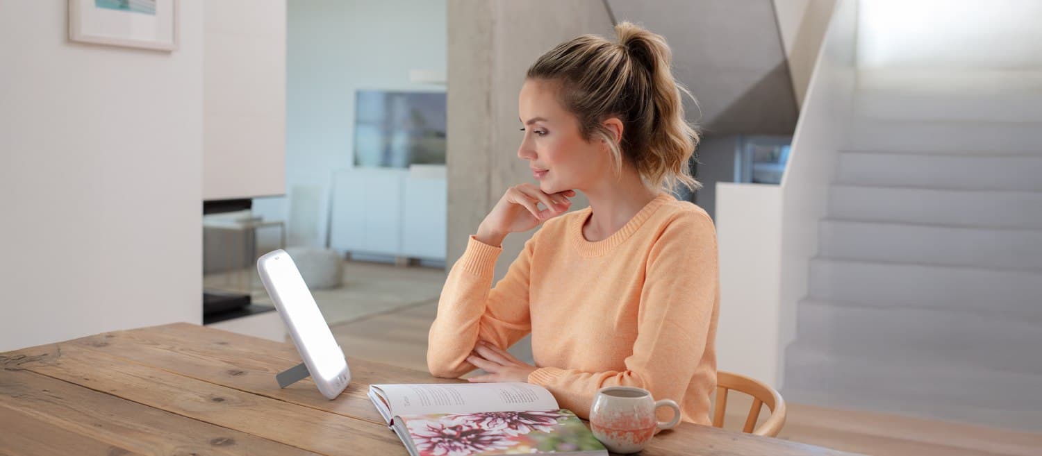 Junge Frau sitzt an einem Tisch mit eingeschalteter Tageslichtlampe, liest ein Buch, daneben steht eine Tasse.