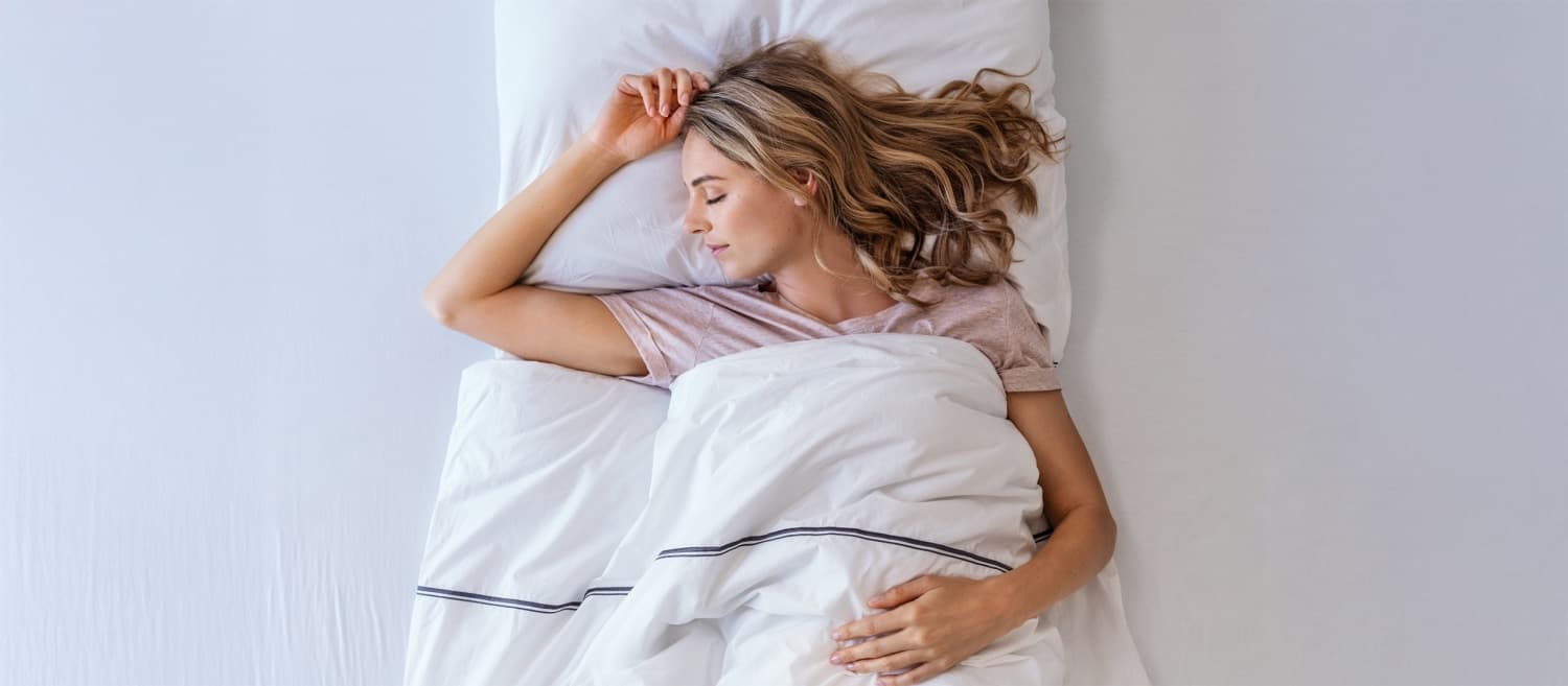 Giovane donna sdraiata su un letto, coperta con una coperta bianca, e indossa una maglietta chiara. Una mano è sul cuscino, l'altra sulla coperta.