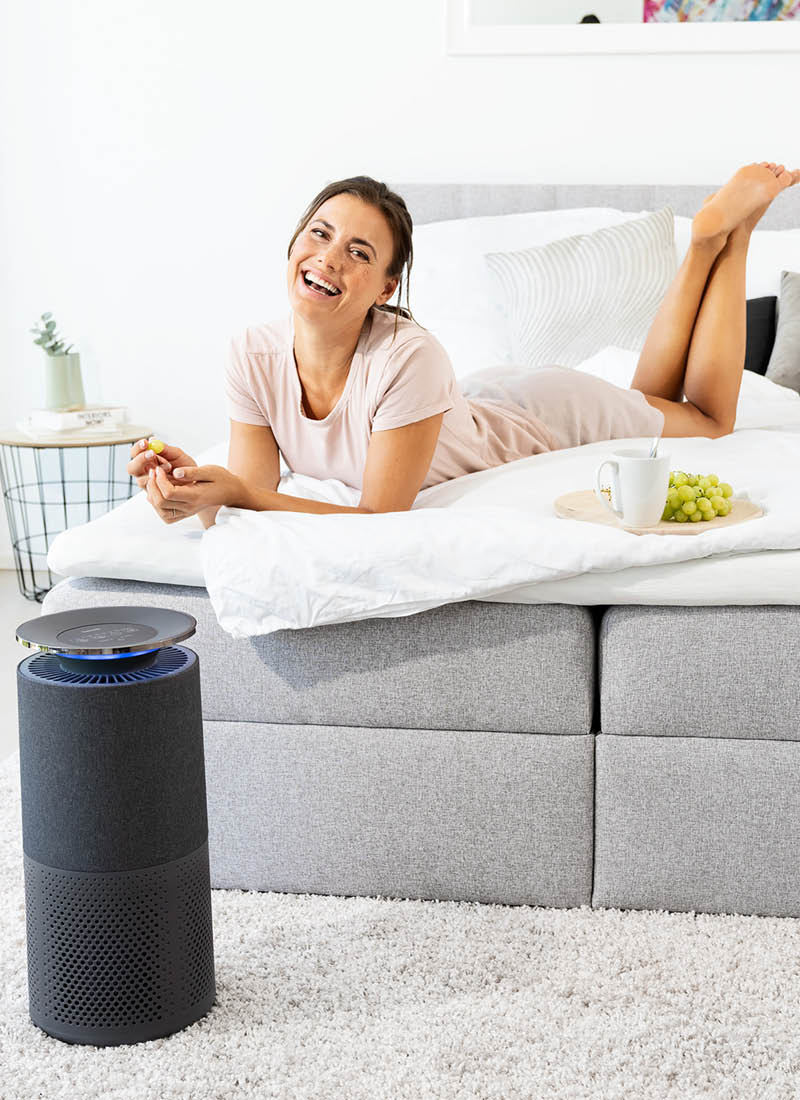 Jeune femme allongée en souriant dans le lit et le purificateur d'air Beurer LR 401 est au premier plan
