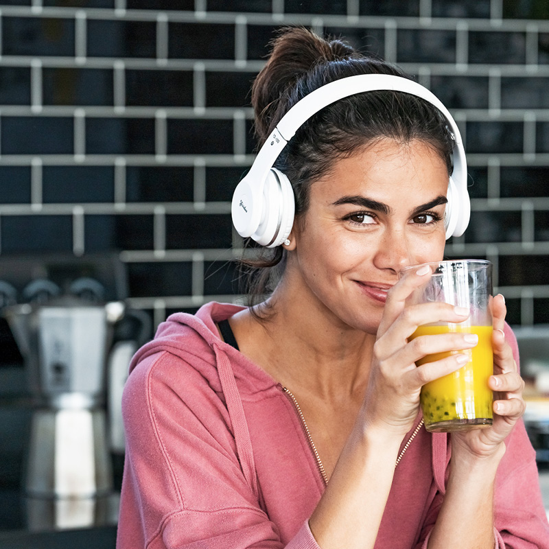 Une femme avec des écouteurs profite d'un verre de jus dans la cuisine et sourit détendue à la caméra