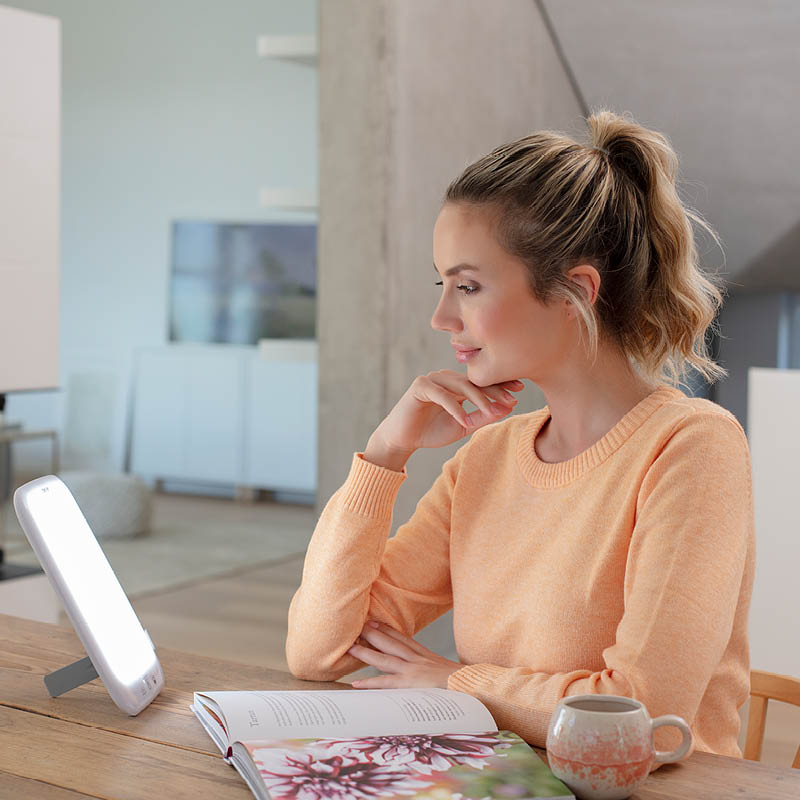 Jeune femme lit un livre à table tout en utilisant la lampe de luminothérapie Beurer TL 85