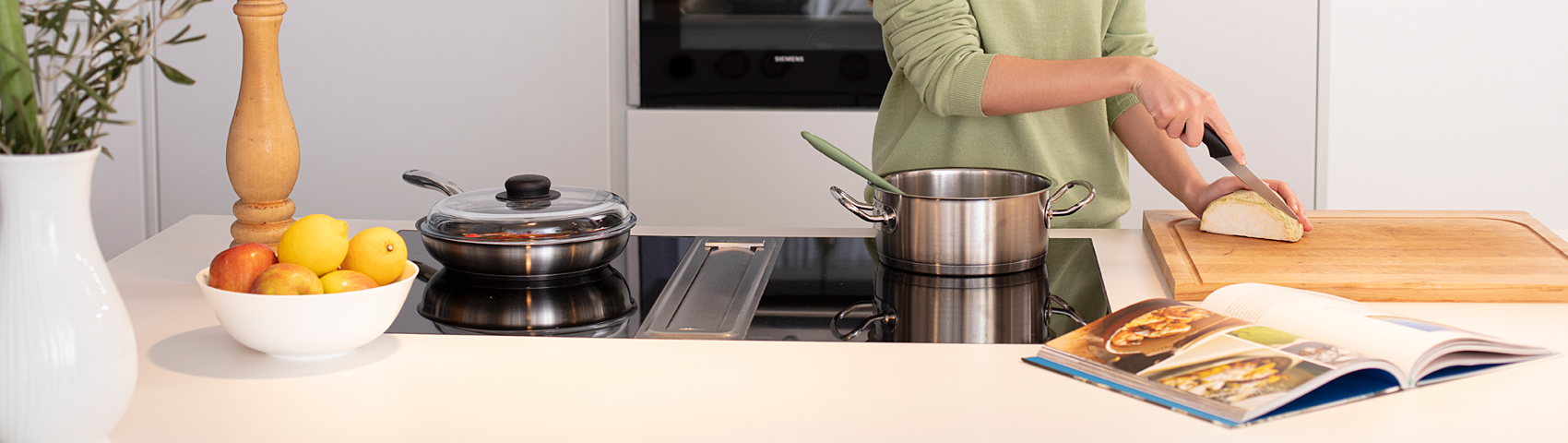 Femme souriante derrière une cuisinière coupe des légumes sur une planche et prépare un repas avec un livre de cuisine