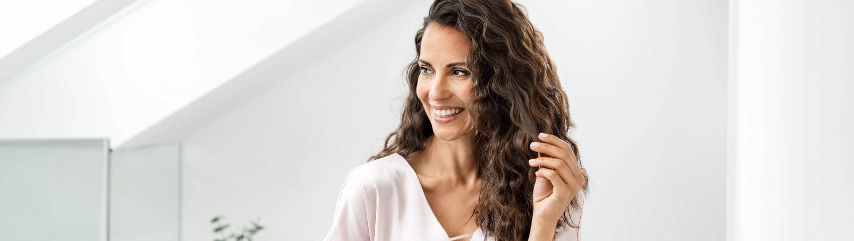 Mujer con cabello largo, castaño y rizado está sonriendo en el baño y se pasa la mano por el cabello