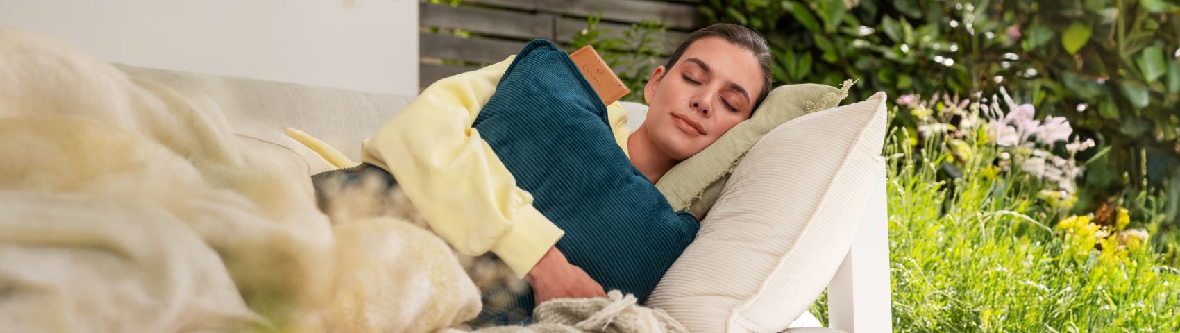 Jeune femme allongée les yeux fermés sur le canapé dans le jardin tenant le coussin chauffant électrique HK 77 de Beurer