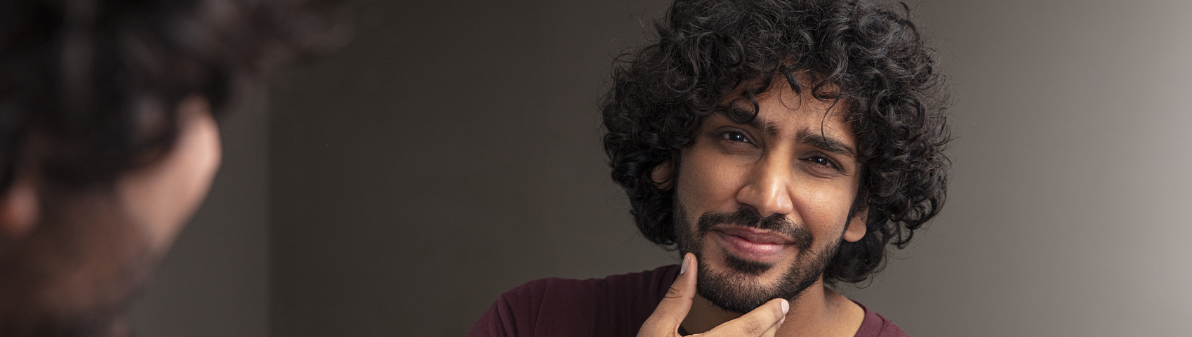 Hombre con cabello oscuro y rizado y barba completa se mira en el espejo y se sostiene la barbilla