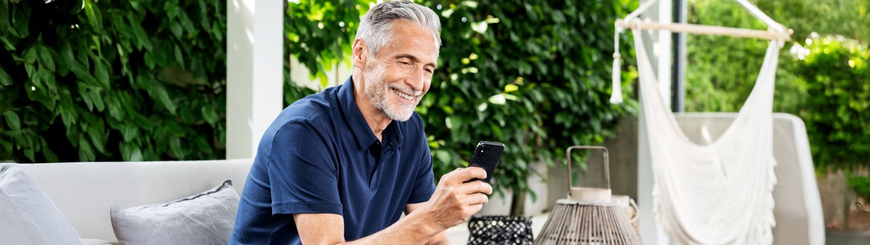 Homme âgé aux cheveux gris et à la barbe assis dehors sur un canapé d'extérieur regardant son téléphone