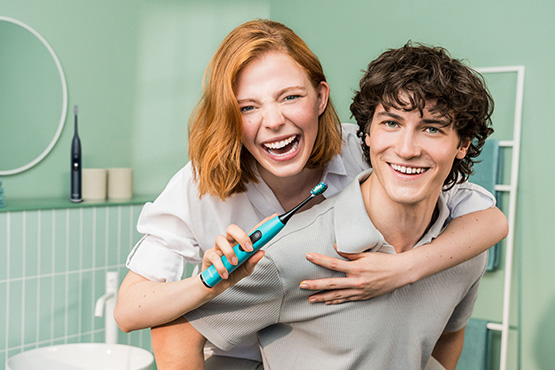 A young man carries a young woman piggyback. She holds a Beurer sonic toothbrush and smiles. A young man carries a young woman piggyback. She holds a Beurer sonic toothbrush and smiles.